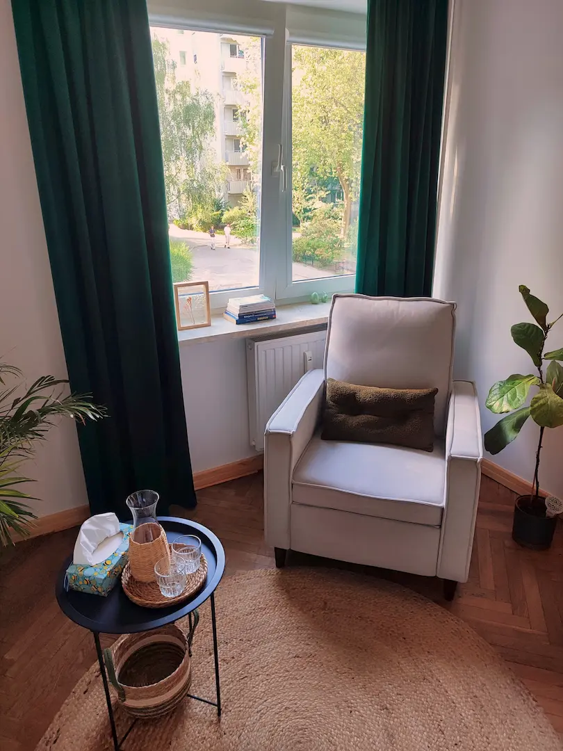Jasny pokój z dużym oknem, zielonymi zasłonami, jasnym fotelem i okrągłym stolikiem na drewnianej podłodze.