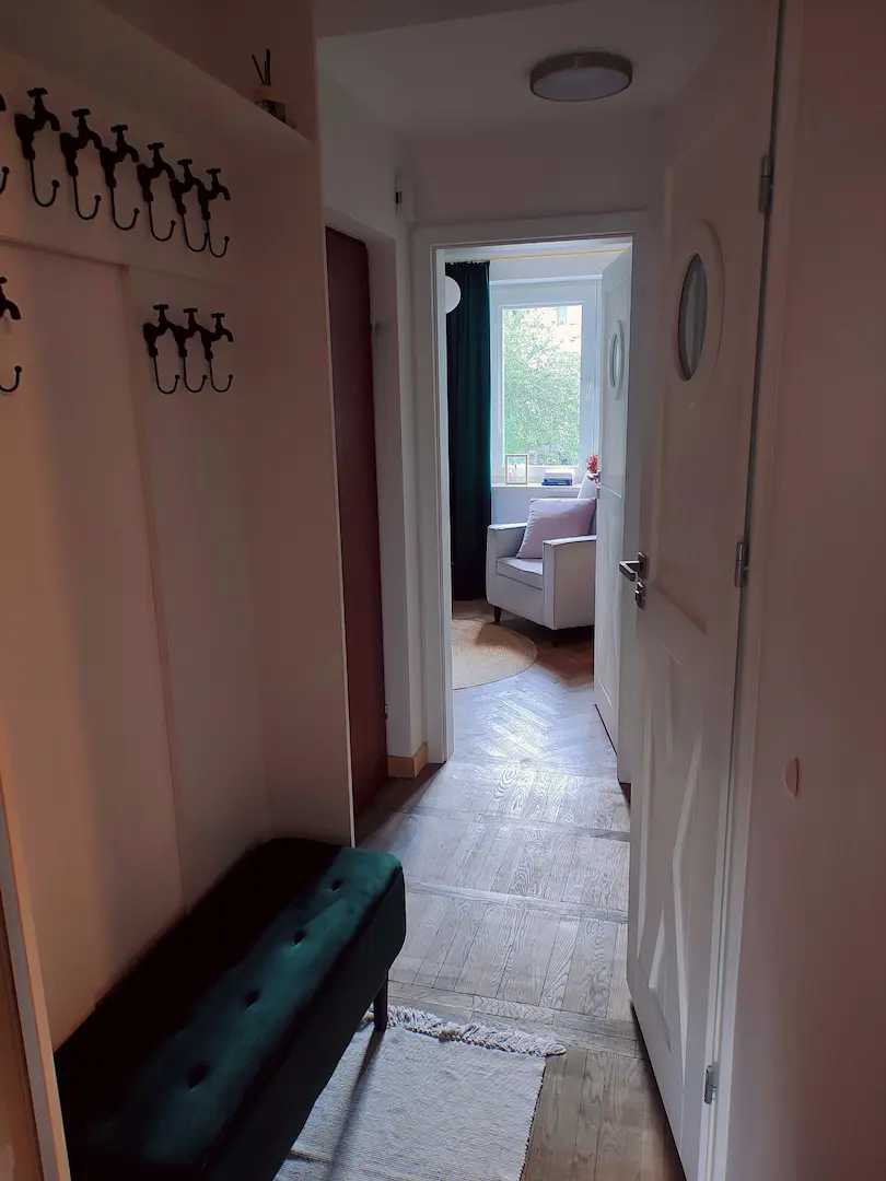 Widok z przedpokoju wzdłuż długiego korytarza z ciemną ławką na buty i otwartymi drzwiami do pokoju.